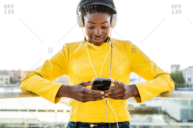 African American woman in stylish bright jacket using mobile phone and listen to music on headphones