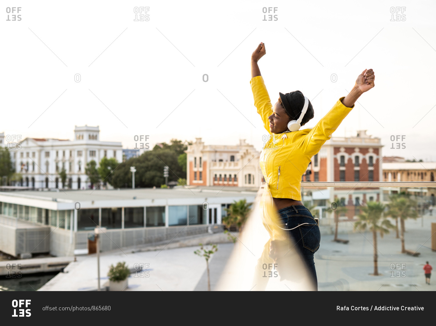 Side view content African American woman in trendy jacket listening to music in headphones while leaning on glass balcony and looking away with arms up on blurred background
