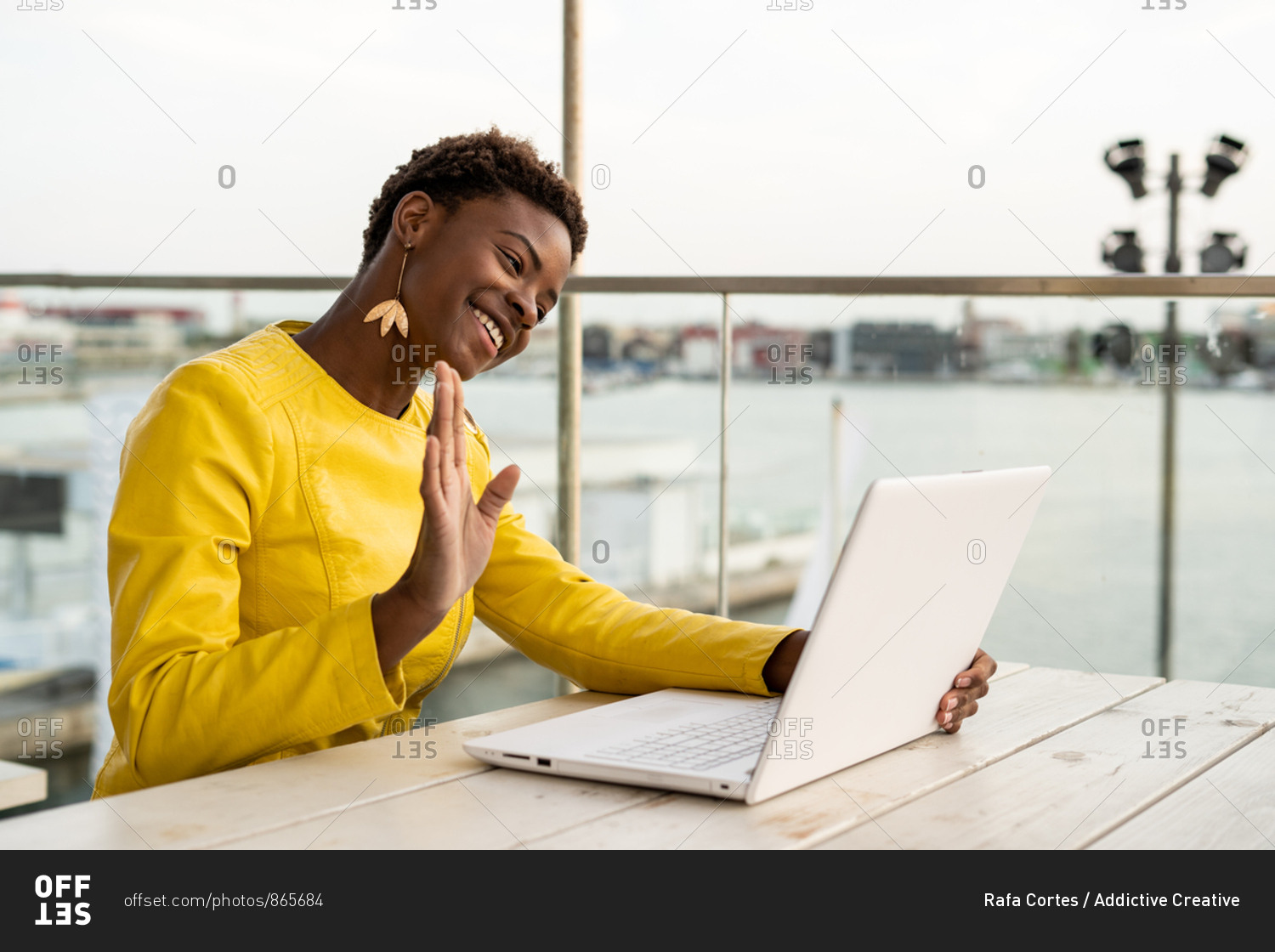 Black African American woman in yellow jacket using laptop web cam at wooden desk in city on blurred background