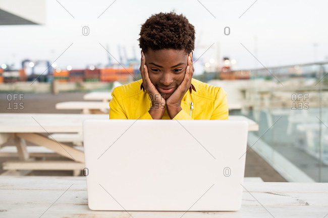 Surprised face of black African American woman in yellow jacket using laptop at wooden desk in city on blurred background