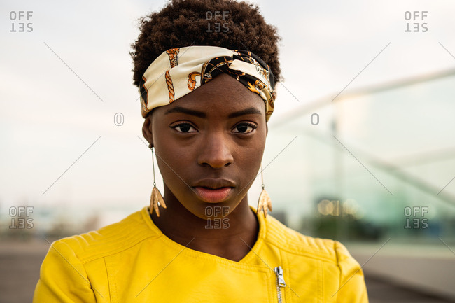 Stylish African American female in modern jacket relaxing sitting on wooden floor and looking at camera