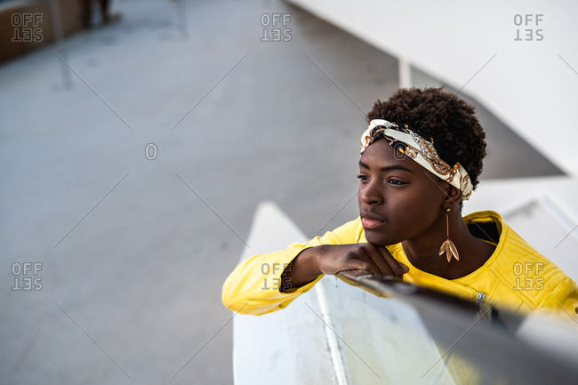 High angle of glad African American woman in stylish wear chilling on stairs leaning on the handrail and looking away