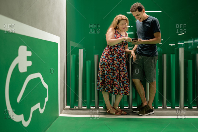 Smiling joyful couple waiting for electric car to charge and sharing mobile phone while standing in light hall