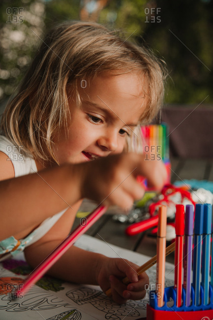 Focused little girl leaning on table and coloring pictures at book with marker pen on blurred background of room at home