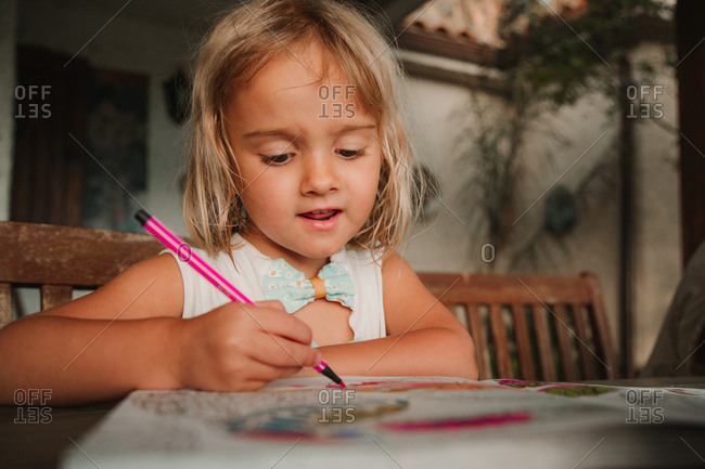 Focused little girl leaning on table and coloring pictures at book with marker pen on blurred background of room at home