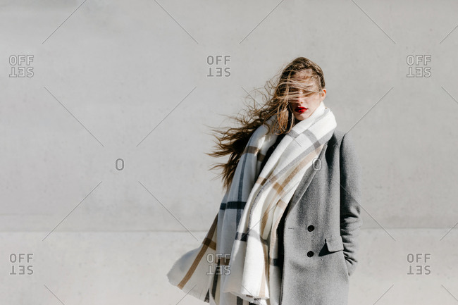 Young female with closed eyes and in stylish gray coat and warm scarf standing against building wall on city street on windy day
