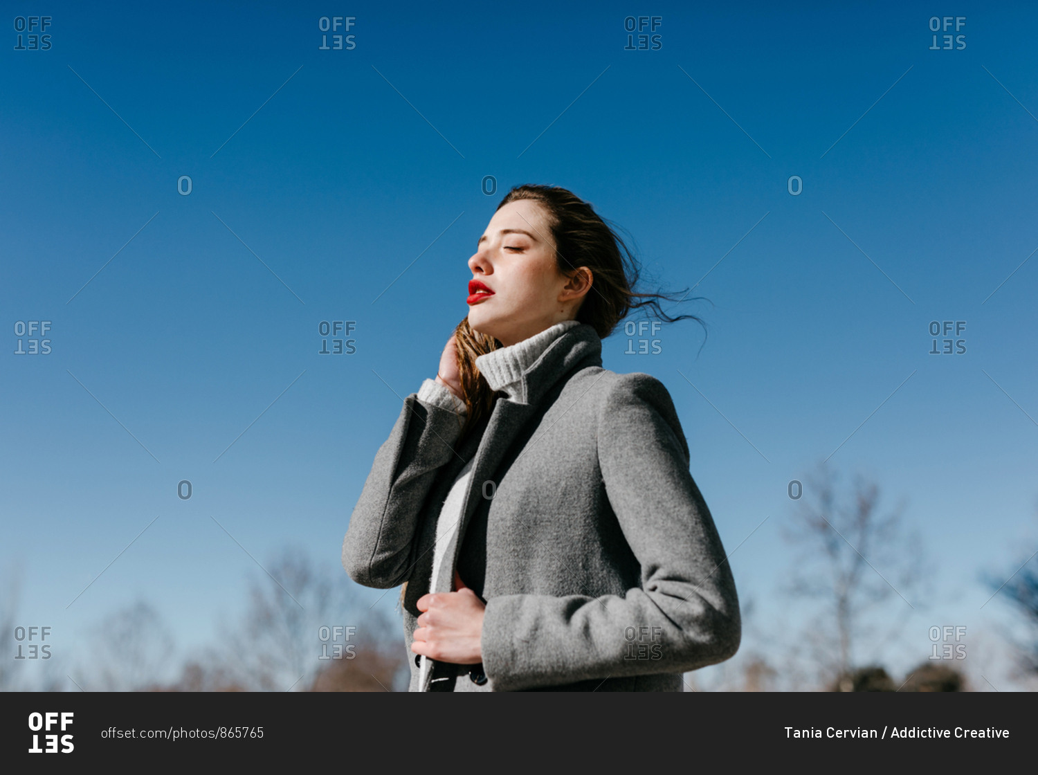 Side view of young female with closed eyes and in stylish gray warm coat standing against clear blue sky on windy day