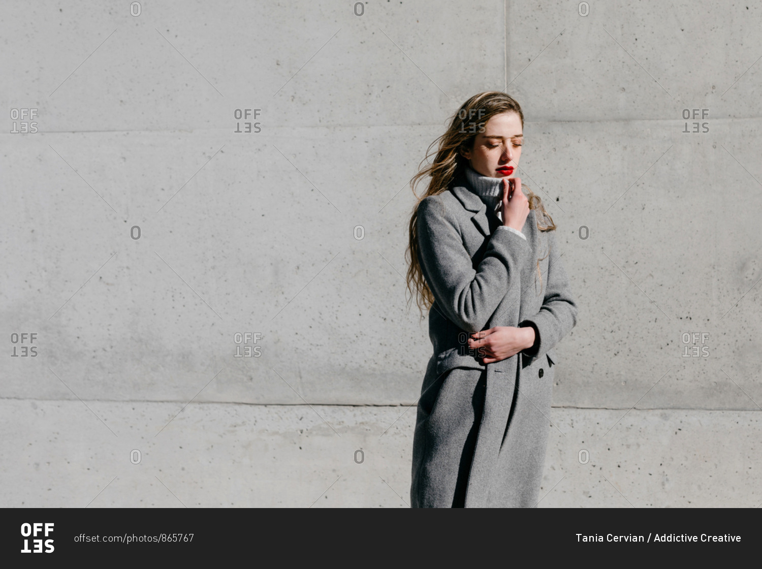 Side view of young female with closed eyes and in stylish gray warm coat standing against building wall on city street on windy day