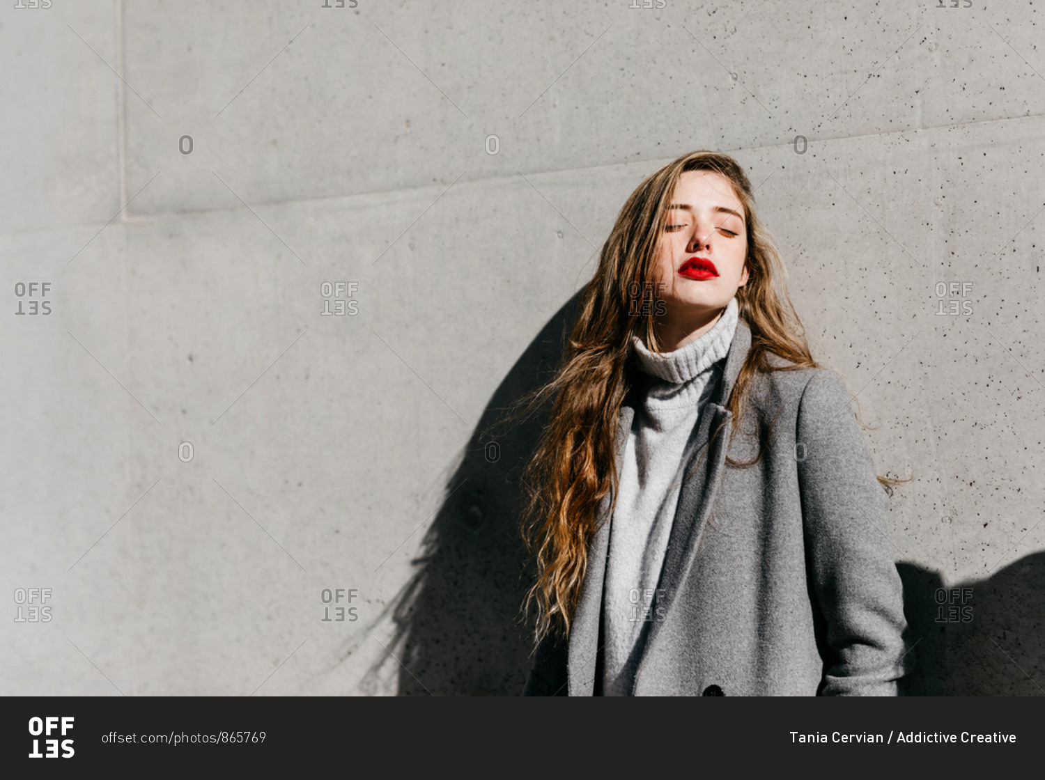 Young female with closed eyes and in stylish gray warm coat standing against building wall on city street on windy day