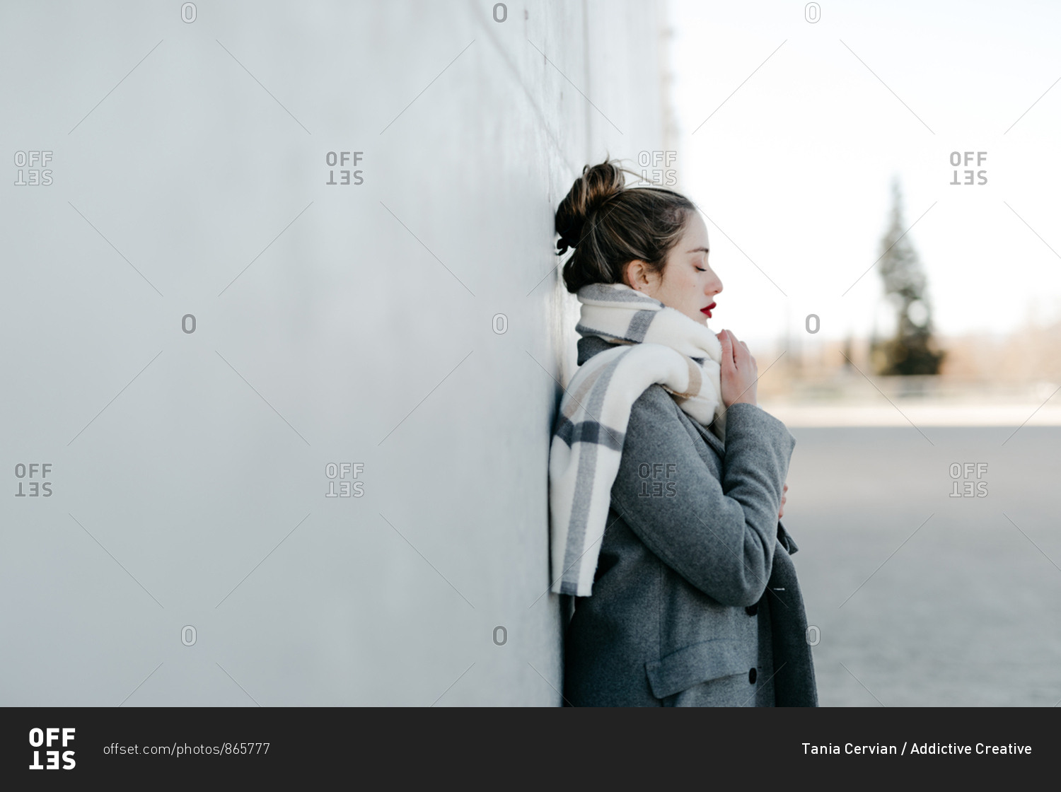 Side view of female with closed eyes and in stylish gray coat and warm scarf leaning against building wall on city street on windy day