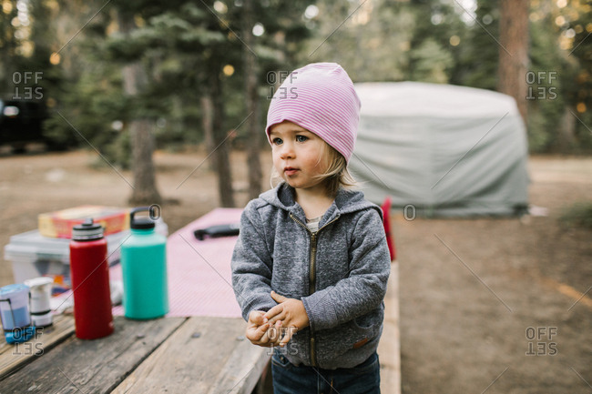 A toddler girl wearing a beanie stands on a picnic table bench in a campground