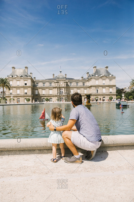 Candid portrait of a father squatting beside his toddler daughter as they watch the boats in the Jardin de Luxembourg in Paris
