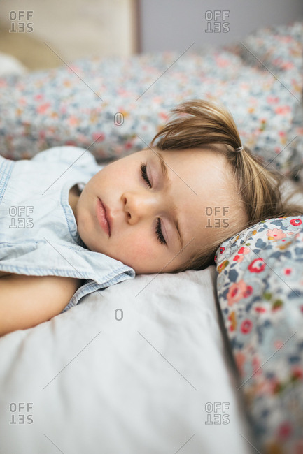 A toddler girl naps on a bed in a bright room surrounded by pillows