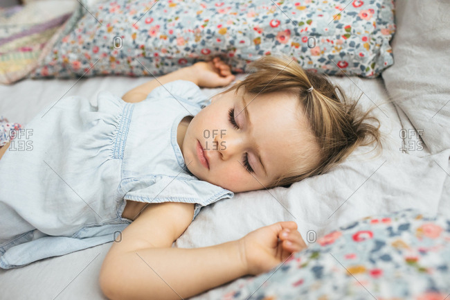 A toddler girl naps on a bed in a bright room surrounded by pillows