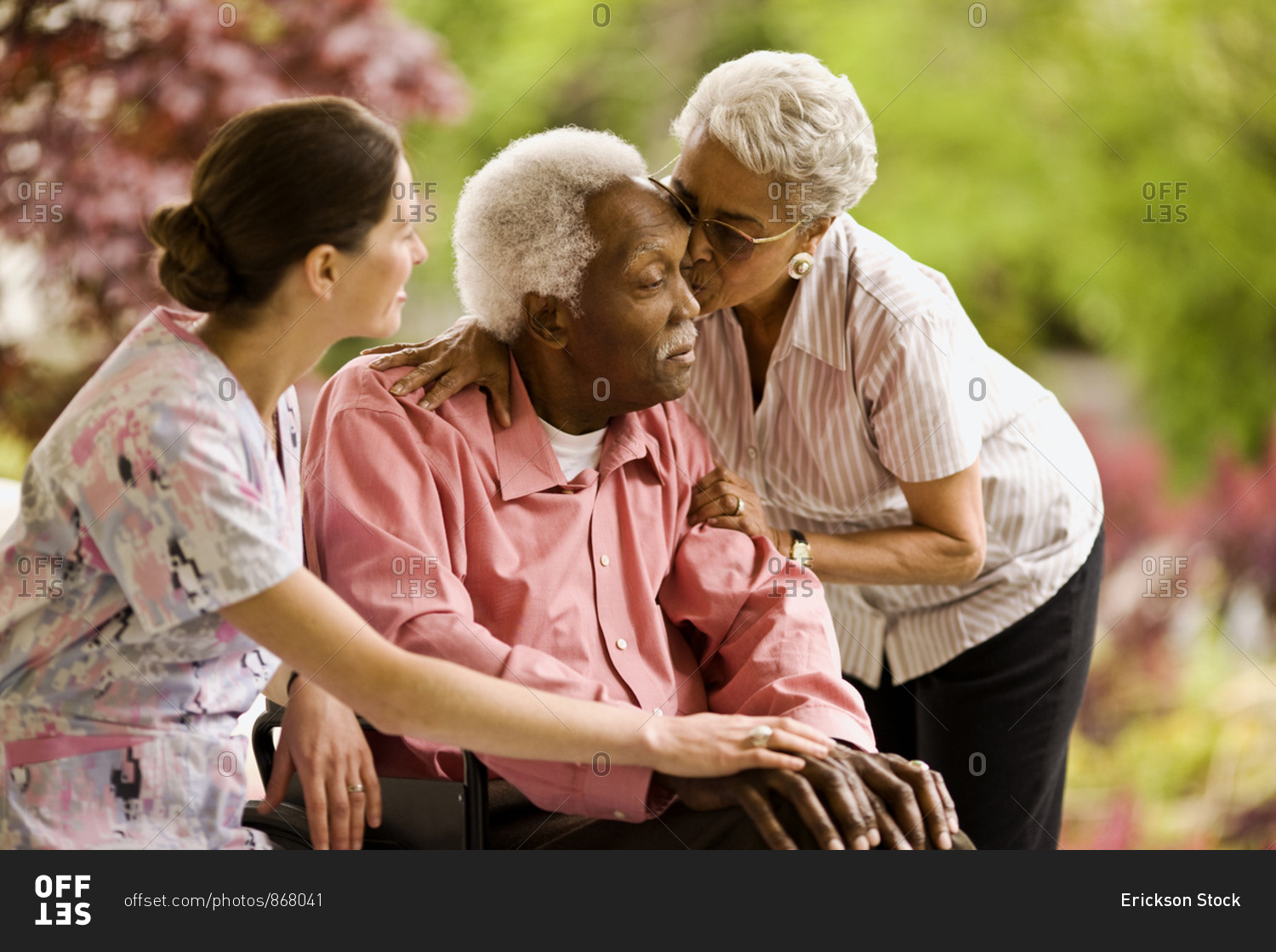 Nurse puts supportive hand on a senior man's knee as a senior woman leans down to hug him and kiss his forehead.