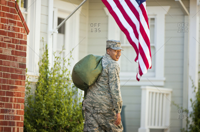 Portrait of a smiling young army soldier carrying a duffel bag outside his home.