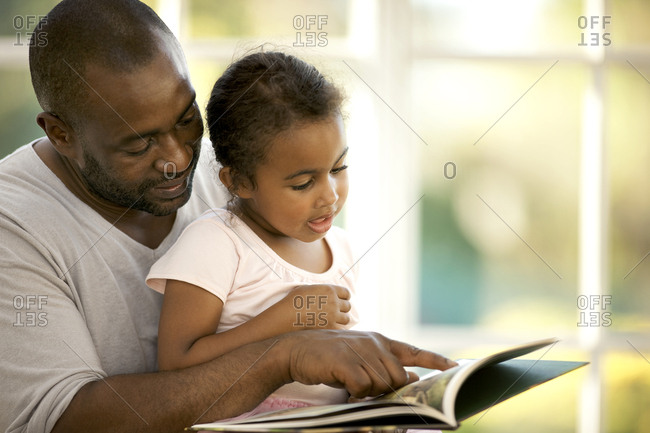 Young girl sitting on her father's lap while reading a book.