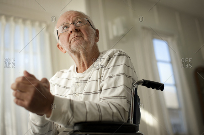 Hopeful senior man looking upward while sitting in a wheel chair with his hands clasped.