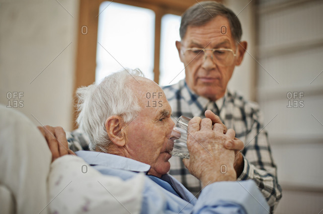 Ailing senior man drinking a glass of water in bed with the help of his concerned male friend.