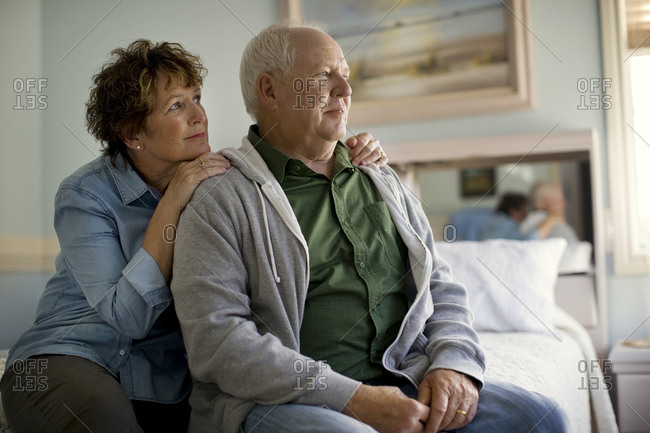 Mature woman lays comforting hands on her husband's shoulders as he stares gloomily out of the bedroom window.