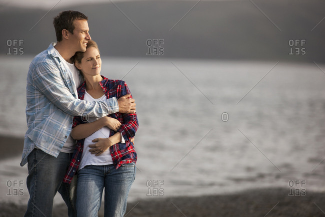 Middle-aged man gently embraces his pregnant wife on the beach.