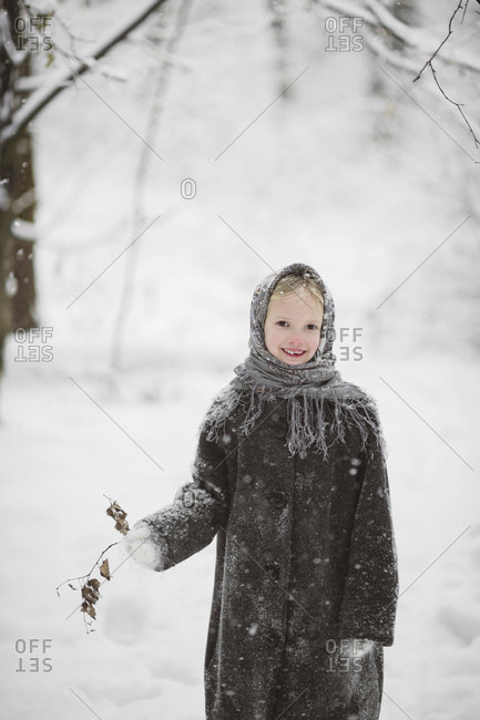 Portrait of little girl wearing headscarf and coat standing in winter forest holding twig with autumn leaves