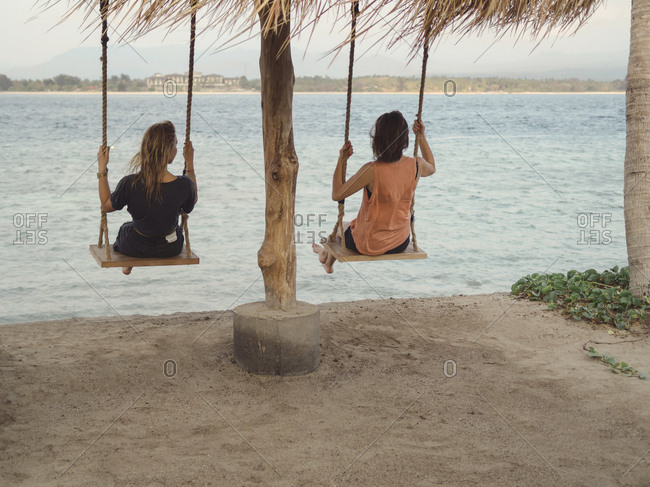 Back view of two women sitting on swings at seafront- Gili Islands- Bali