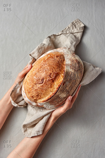 Homemade freshly baked loaf of bread from whole grain flour on a textile towel in the woman's hands on a gray background with copy space.