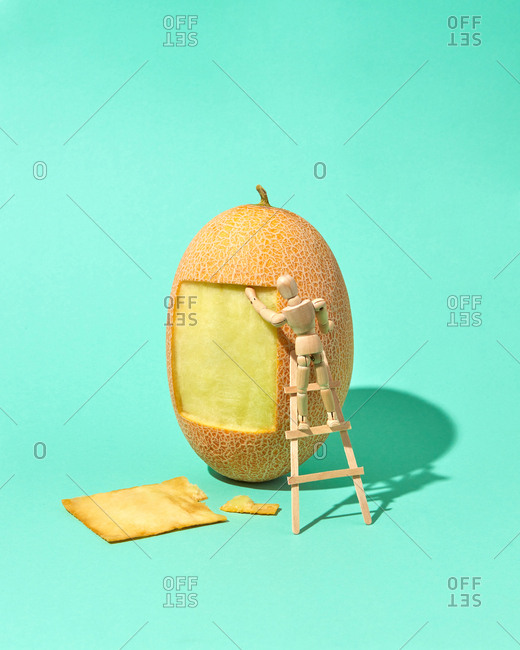 Square part of melon's peel is cut out by miniature articulation mannequin on a wooden ladder with hard shadows on a biscay green background, place for text.
