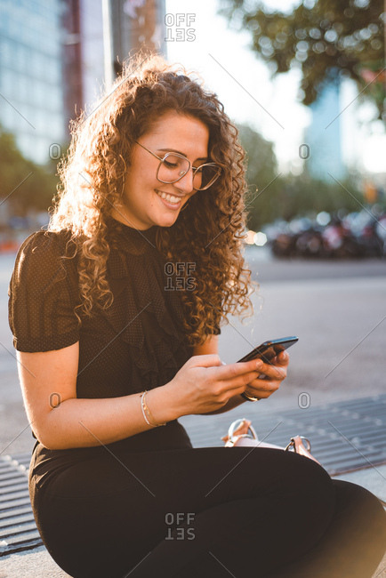 Woman with curly hair sitting on streetside using cell phone in Barcelona in evening light