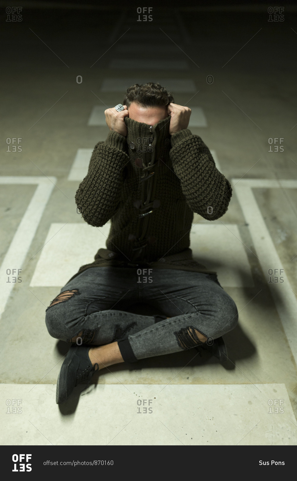 Portrait of a man sitting in a parking garage at night covering his face with sweater