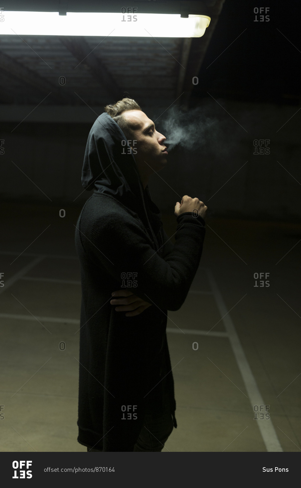 Profile view of a man in a parking garage at night covering his face with sweater
