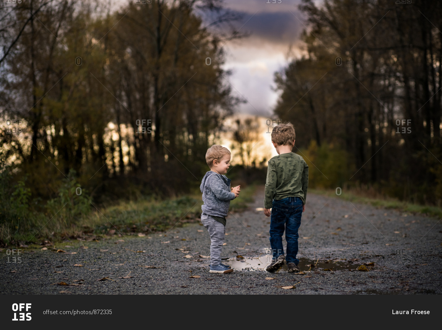 Rear view of two little boys walking on dirt path with puddles at sunset
