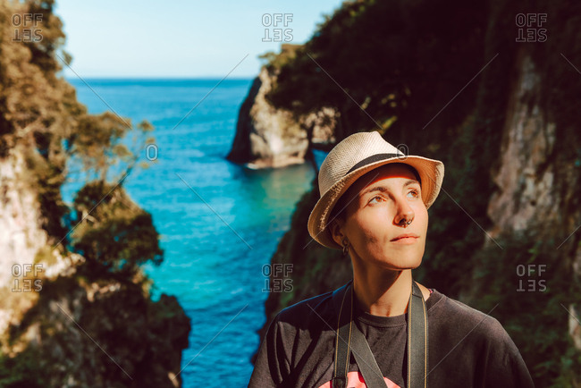 Young woman in hat standing with camera hanging on neck and enjoying picturesque view of sea and rocks in Ribadedeva Asturias Spain