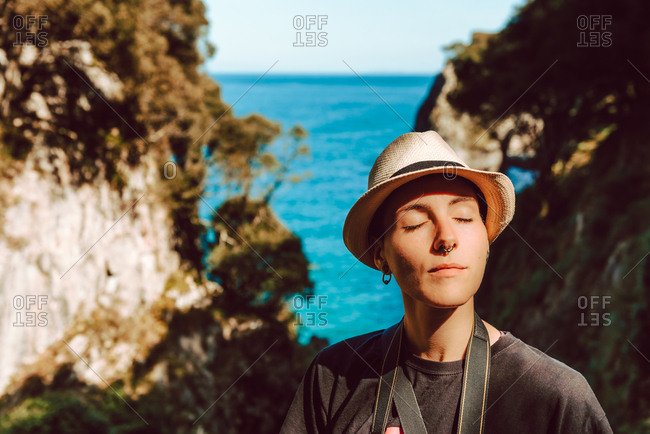 Young woman in hat standing with camera hanging on neck and enjoying picturesque view of sea and rocks in Ribadedeva Asturias Spain