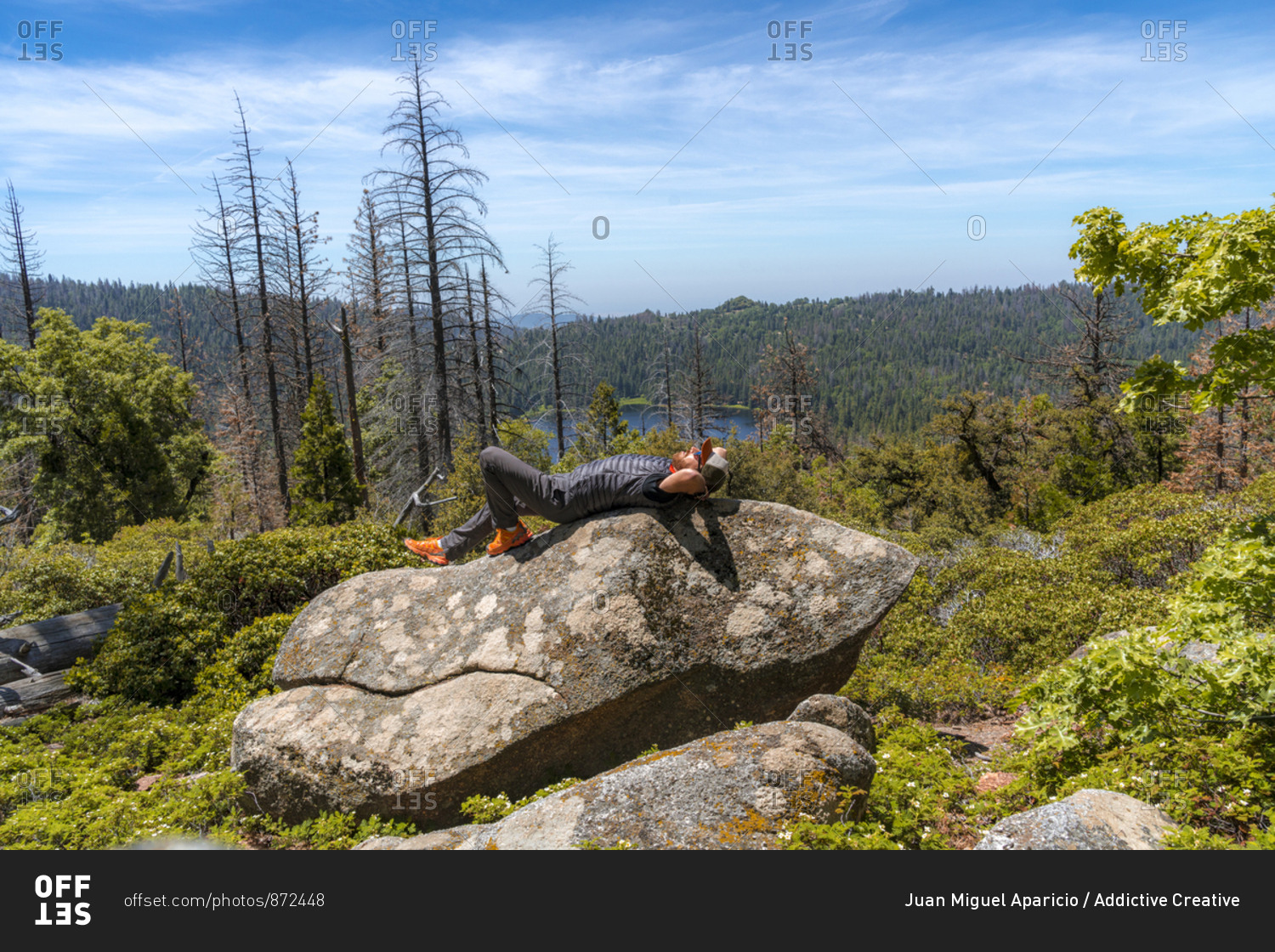 Side view of carefree man in cap lying on back with hands behind head and looking up on stone surrounded with greenery in USA