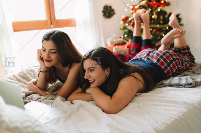 Two women friends in winter holidays at home looking something on laptop and mobile phone near Christmas tree in cozy interior. Interior with Christmas decorations.