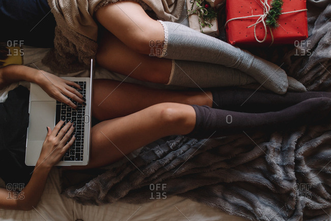 Two friends with knee high socks in winter holidays at home writing something on laptop near Christmas tree in cozy interior with Christmas presents.