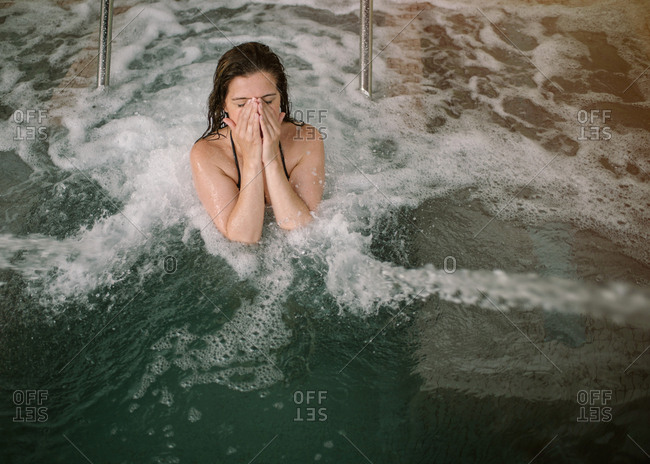 From above adult female with closed eyes in bikini covering face from water while swimming in pool with jets and bubbles