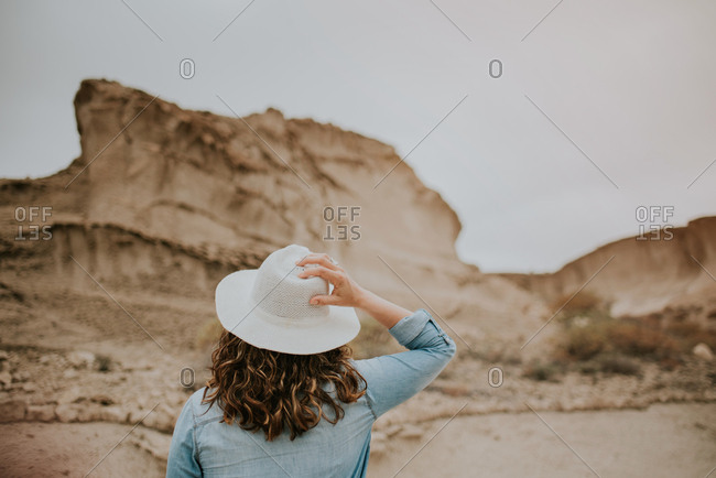 Back view of unrecognizable woman in casual clothes holding hat in her head while walking on desert sandy dunes on gloomy day
