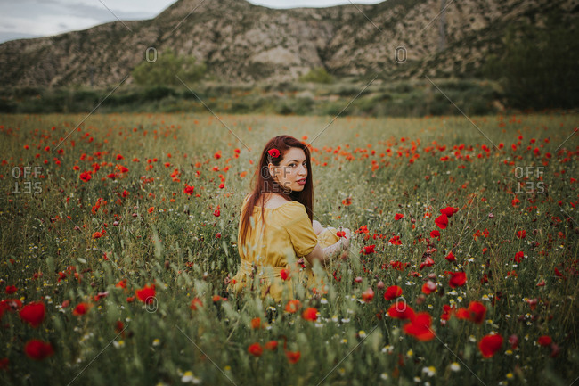 Pensive attractive red haired adult lady in yellow dress with red poppy in hair and red lips looking over shoulder