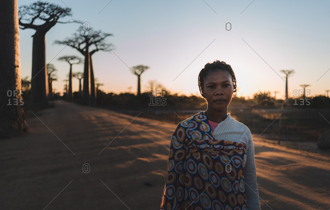 Madagascar - JULY 6, 2019: Confident ethnic woman in exotic multi colored outfit standing at slope by tall baobab trees in sun rays at twilight