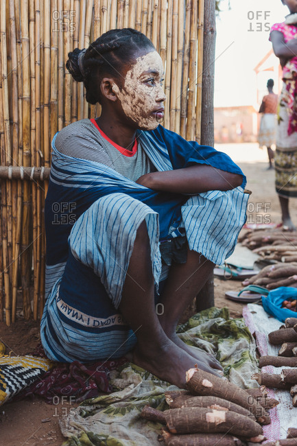Madagascar - JULY 6, 2019: Exotic ethnic person in multi colored modern outfit sitting by fence and selling piles of edible root vegetable