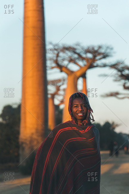Madagascar - JULY 6, 2019: Content African woman in exotic multi colored cover with hood standing by baobab trees in sun rays at twilight
