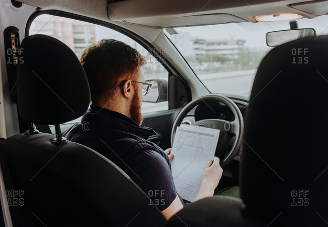 Back view of attentive thoughtful adult man focusing and checking documents while sitting behind steering wheel in car cabin during daytime on blurred background