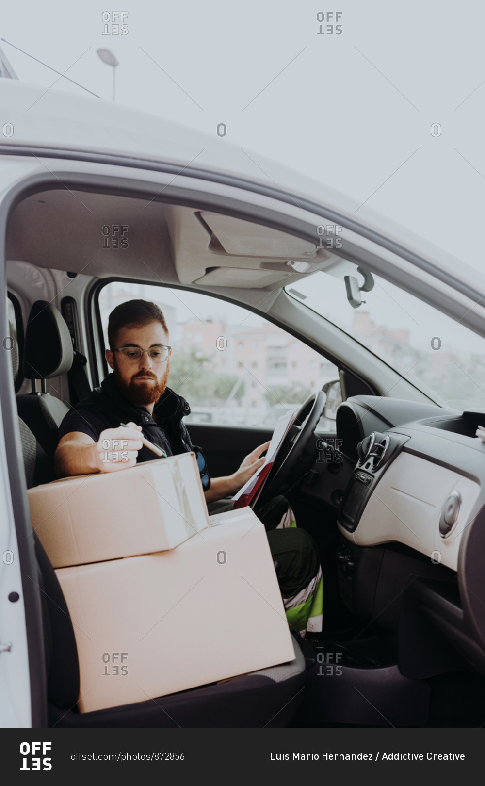 Attentive adult bearded courier in glasses preparing packages for transportation while sitting and marking boxes in car on blurred background during daytime