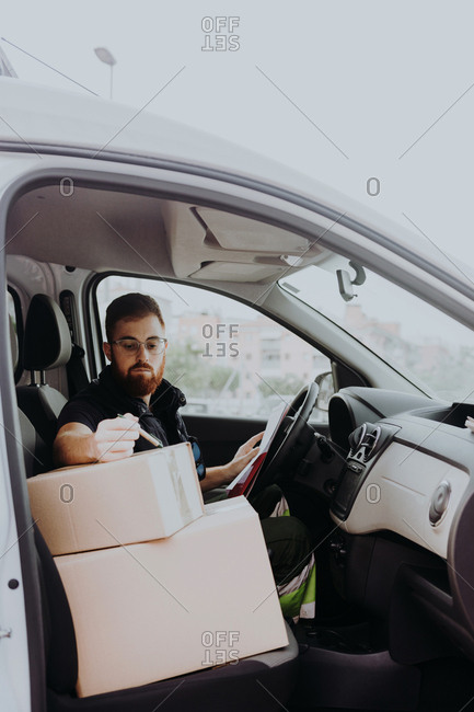Attentive adult bearded courier in glasses preparing packages for transportation while sitting and marking boxes in car on blurred background during daytime