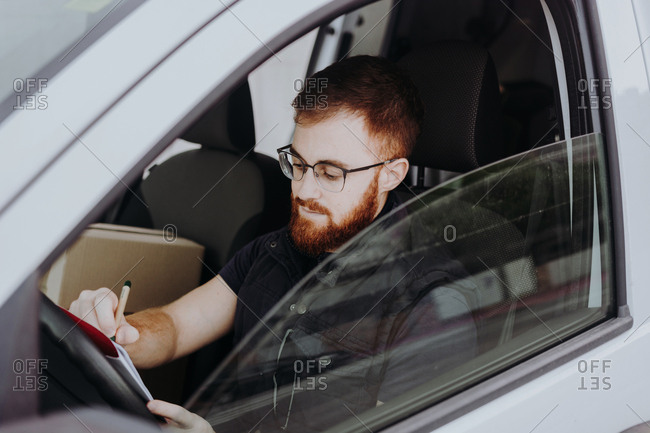 Side view of attentive thoughtful adult man focusing and checking documents while sitting behind steering wheel in car cabin during daytime on blurred background