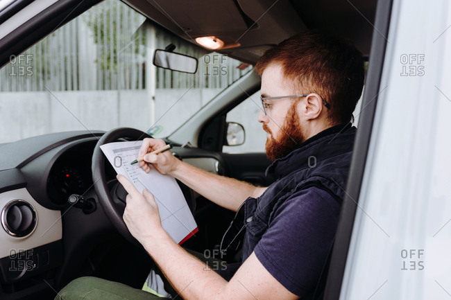 Side view of attentive thoughtful adult man focusing and checking documents while sitting behind steering wheel in car cabin during daytime on blurred background
