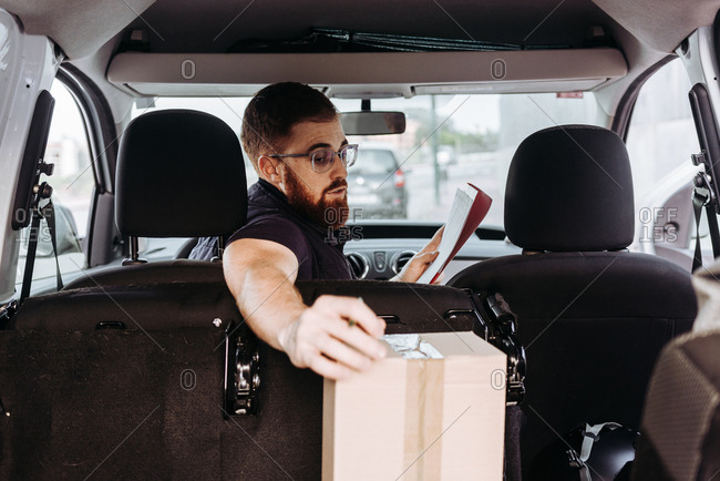 Attentive adult bearded courier in glasses preparing packages for transportation while sitting and marking boxes in car on blurred background during daytime